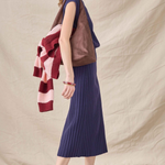 A woman stands in profile wearing a sleeveless brown leather vest over a navy blue pleated midi skirt, with a pink and burgundy striped knit sweater draped over her shoulder, white socks, and burgundy pointed-toe slingback heels against a beige fabric backdrop.