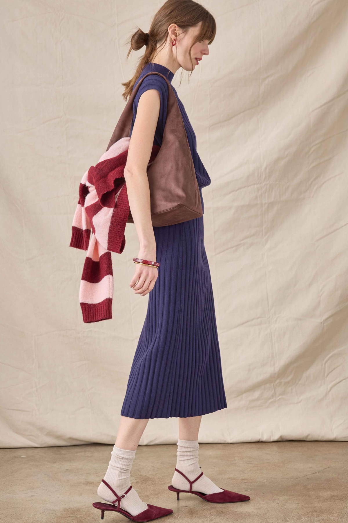 A woman stands in profile wearing a sleeveless brown leather vest over a navy blue pleated midi skirt, with a pink and burgundy striped knit sweater draped over her shoulder, white socks, and burgundy pointed-toe slingback heels against a beige fabric backdrop.