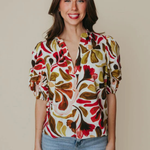 A woman modeling a short-sleeve blouse with a bold abstract floral pattern in red, olive green, mustard yellow, and brown on a cream background, paired with light blue jeans against a solid beige studio backdrop.