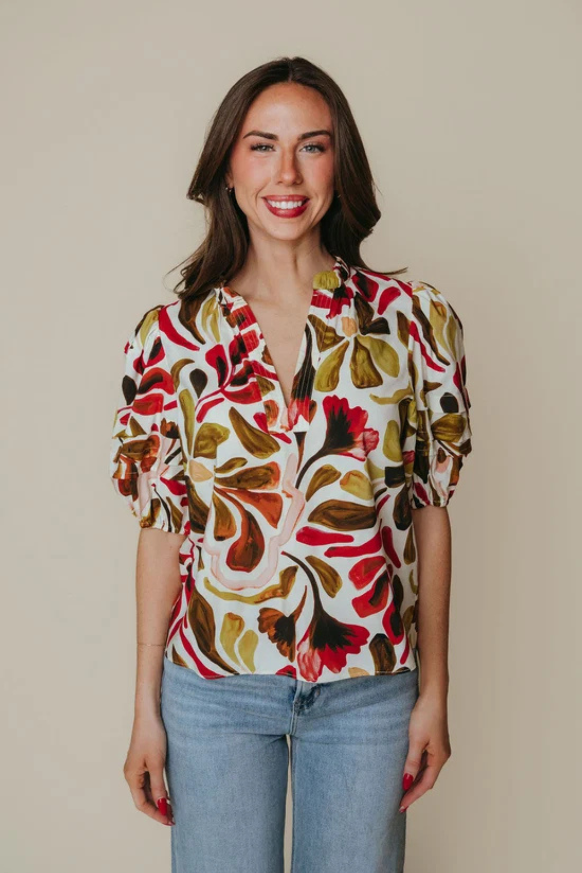 A woman modeling a short-sleeve blouse with a bold abstract floral pattern in red, olive green, mustard yellow, and brown on a cream background, paired with light blue jeans against a solid beige studio backdrop.