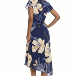 A woman modeling a navy blue midi dress with a large cream and gray floral print, shown from a side-back angle against a plain white background.