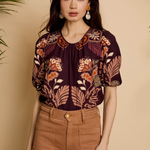 A woman modeling a short-sleeve blouse with a dark purple base and a warm-toned floral and leaf print in orange, beige, and brown, paired with brown pants against a beige background.