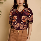A woman modeling a short-sleeve blouse with a dark purple base and a warm-toned floral and leaf print in orange, beige, and brown, paired with brown pants against a beige background.