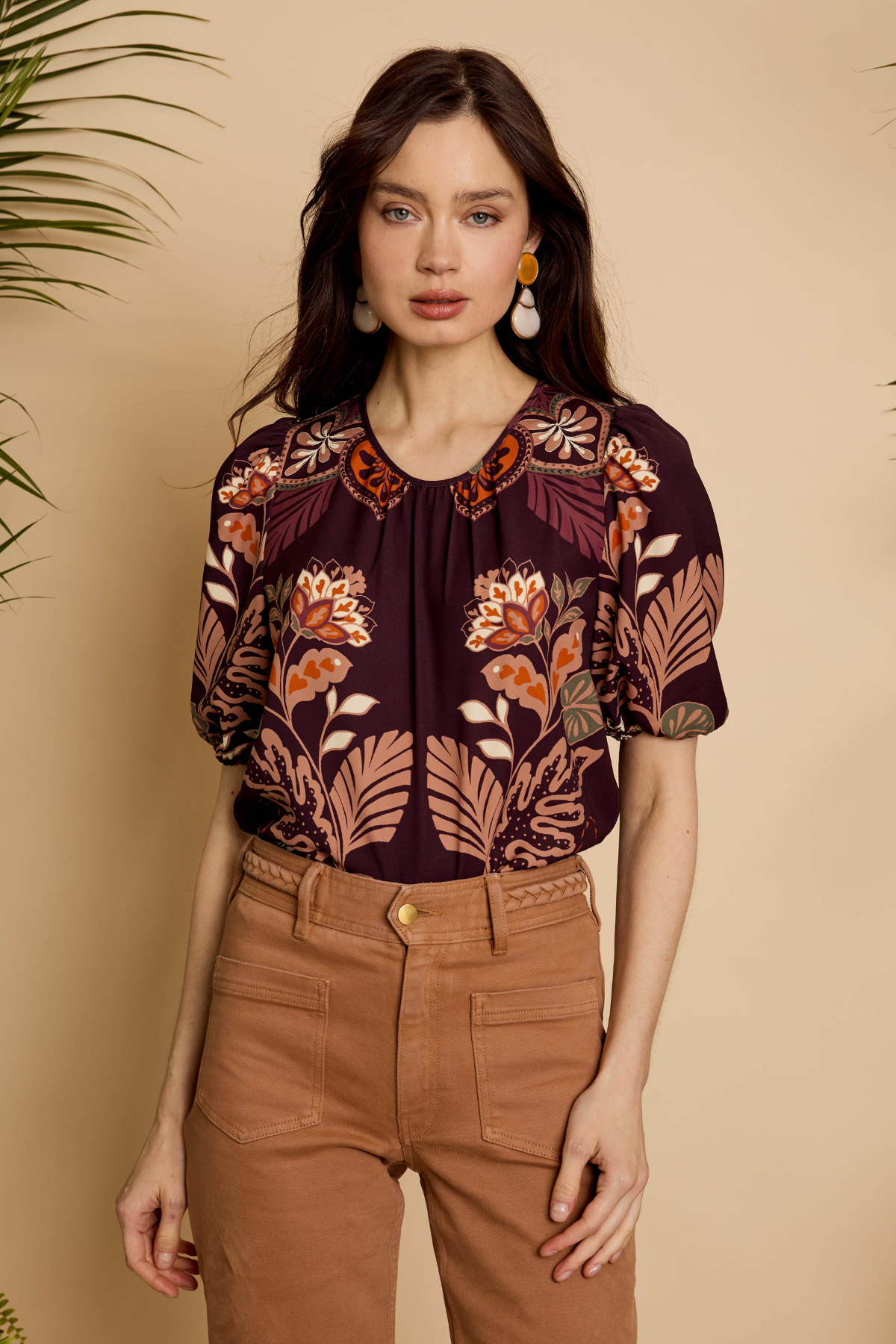 A woman modeling a short-sleeve blouse with a dark purple base and a warm-toned floral and leaf print in orange, beige, and brown, paired with brown pants against a beige background.