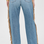 Rear view of a person wearing light blue denim jeans with long tan fringe draped down the outer sides of the legs, against a plain light background.