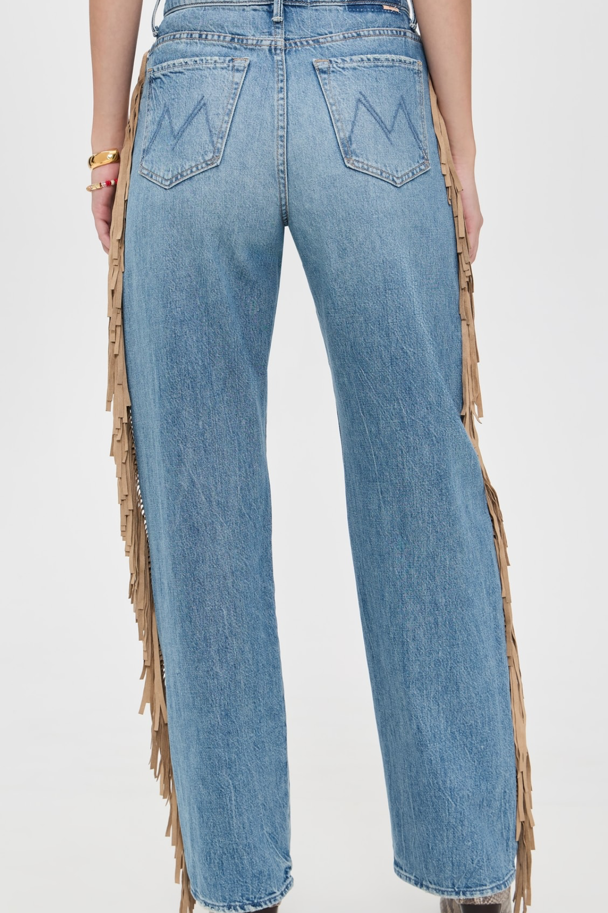 Rear view of a person wearing light blue denim jeans with long tan fringe draped down the outer sides of the legs, against a plain light background.