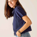 A woman with long brown hair wearing a navy blue short-sleeve button-up shirt and light blue jeans, posing against a plain light beige background.