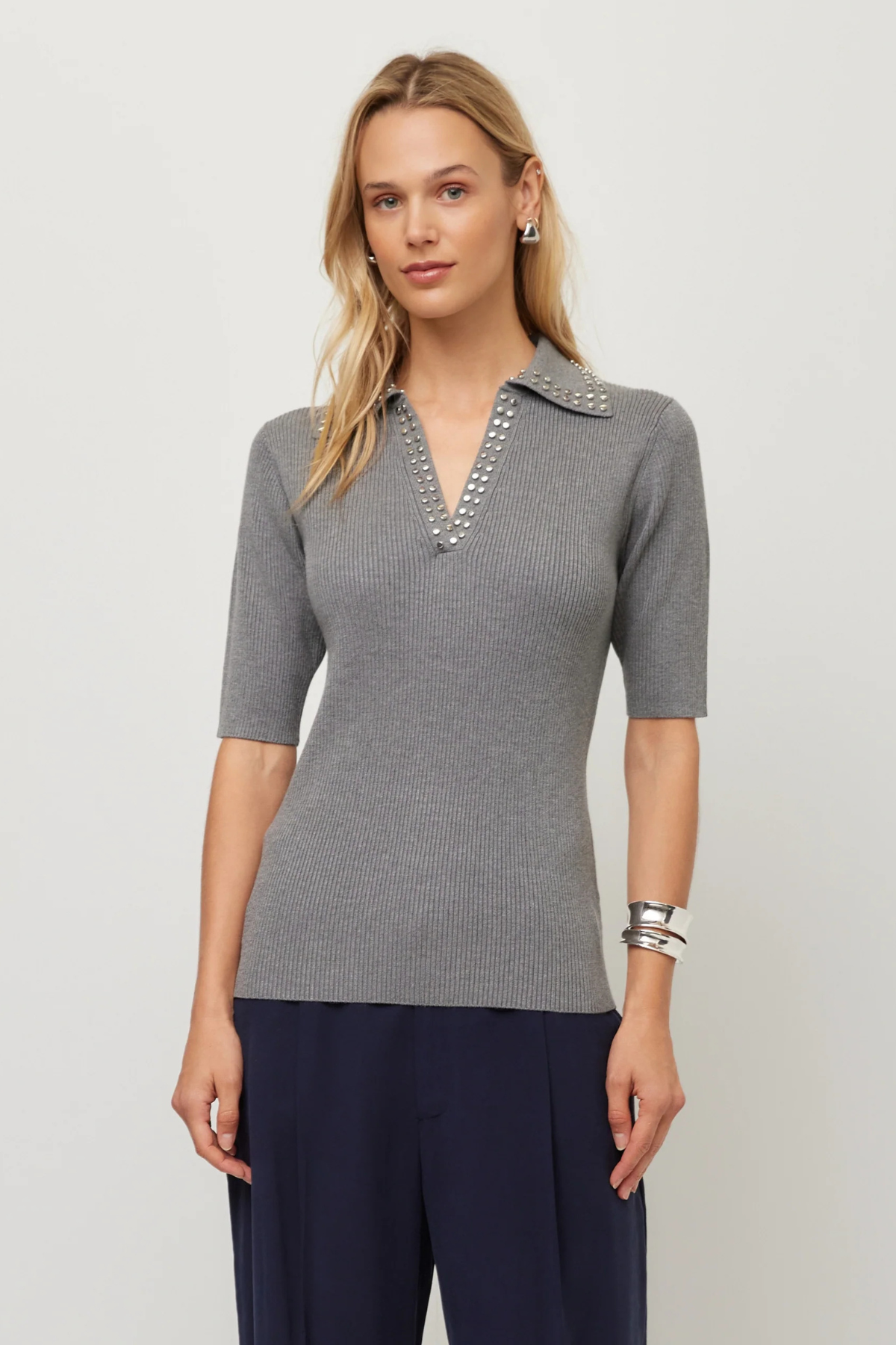 A woman modeling a gray ribbed knit polo shirt with a decorative silver beaded collar, paired with navy blue pants, against a light gray studio background.