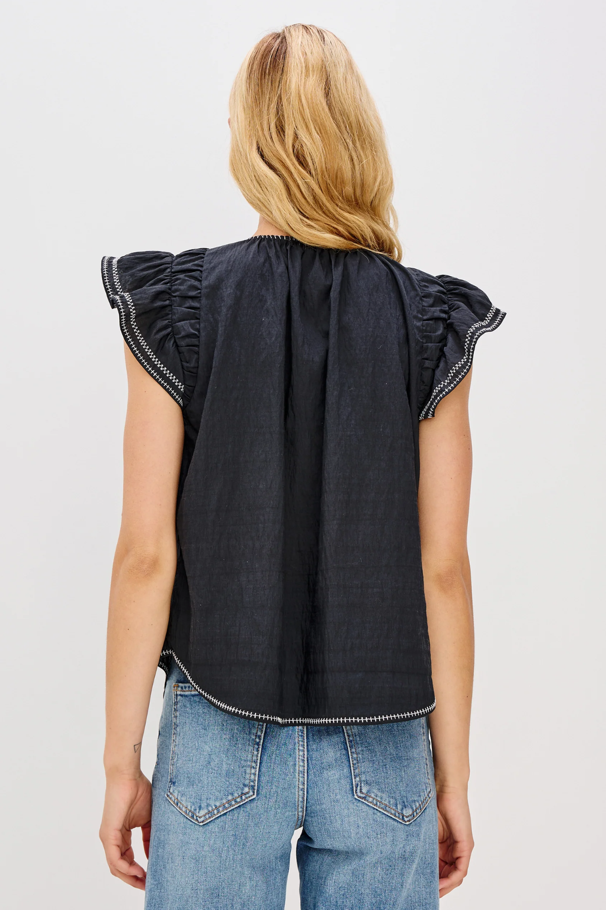 Person wearing a black sleeveless top with ruffled shoulders and blue jeans on a light gray background