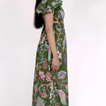 A woman modeling a long green maxi dress with a colorful floral and bird print, shown from a side profile against a plain white studio background.