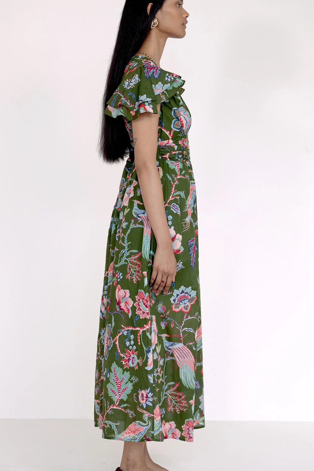 A woman modeling a long green maxi dress with a colorful floral and bird print, shown from a side profile against a plain white studio background.