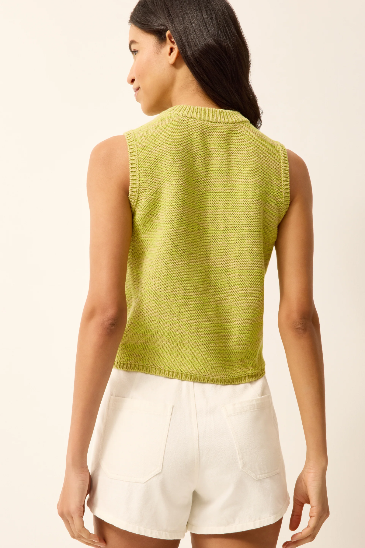 Woman wearing a green sleeveless knitted top and white shorts on a beige background