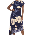 A woman modeling a navy blue midi dress with a large cream and gray floral print, featuring short cap sleeves and an asymmetrical hemline, set against a plain white background.