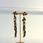 A pair of long dangling earrings displayed on a rose gold stand against a clean white background. The earrings feature metallic gold and dark bronze leaf-like drops with small white pearl accents at the top.