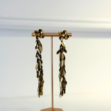 A pair of long dangling earrings displayed on a rose gold stand against a clean white background. The earrings feature metallic gold and dark bronze leaf-like drops with small white pearl accents at the top.