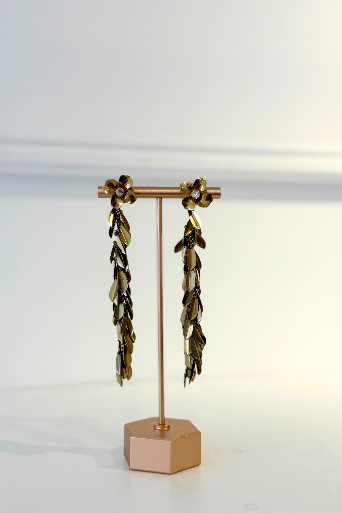 A pair of long dangling earrings displayed on a rose gold stand against a clean white background. The earrings feature metallic gold and dark bronze leaf-like drops with small white pearl accents at the top.