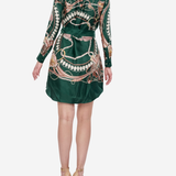 A woman viewed from the back wearing a dark green satin shirt dress with a large gold and white chain print, paired with beige high-heeled sandals, against a light gray background.
