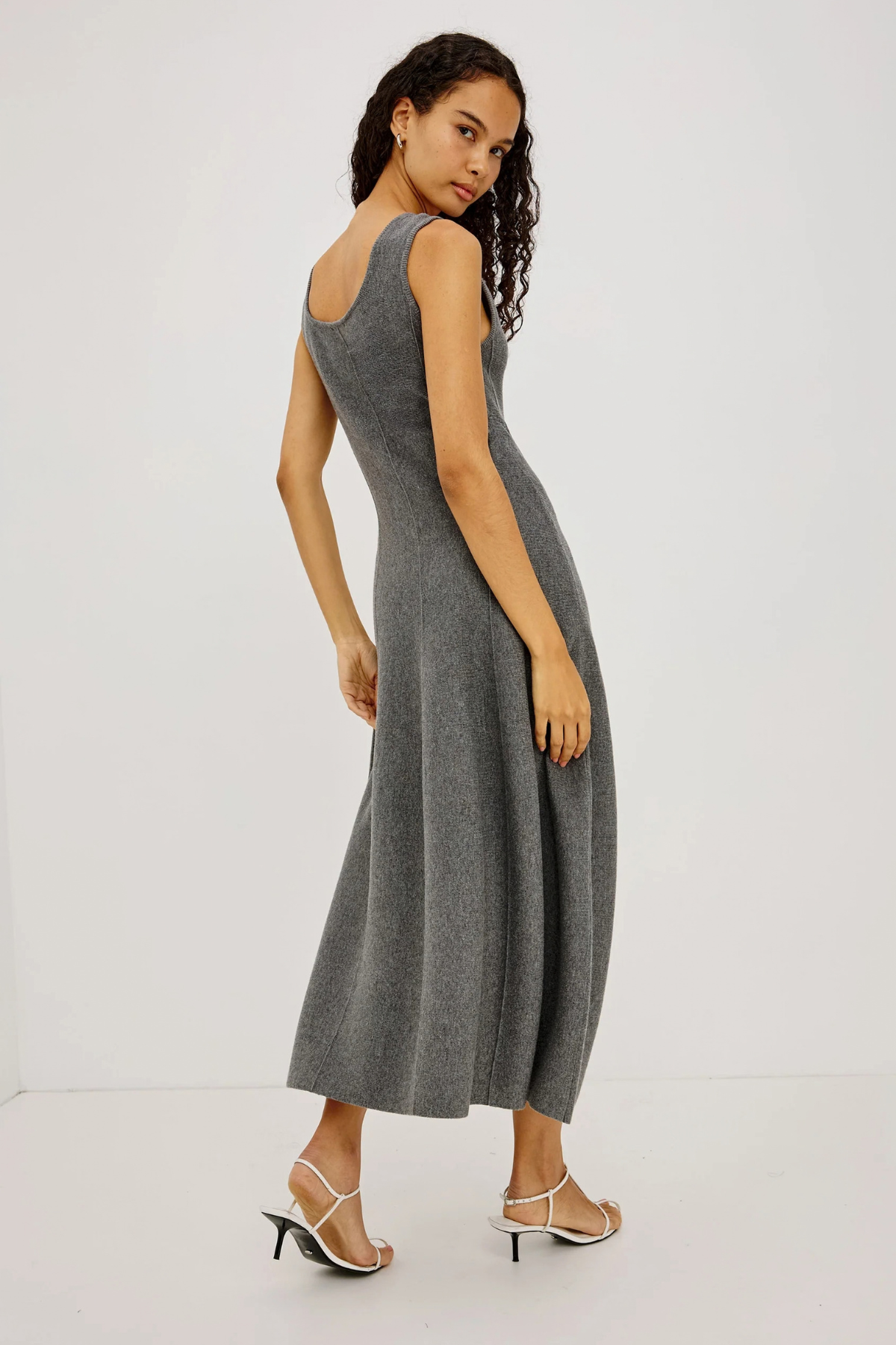 A woman modeling a sleeveless, scoop-neck gray knit midi dress, shown from a three-quarters back angle against a plain white studio background.