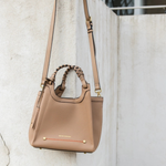 A taupe leather shoulder bag with braided top handles and gold-tone hardware, hanging against a textured concrete wall.