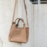 A taupe leather shoulder bag with braided top handles and gold-tone hardware, hanging against a textured concrete wall.