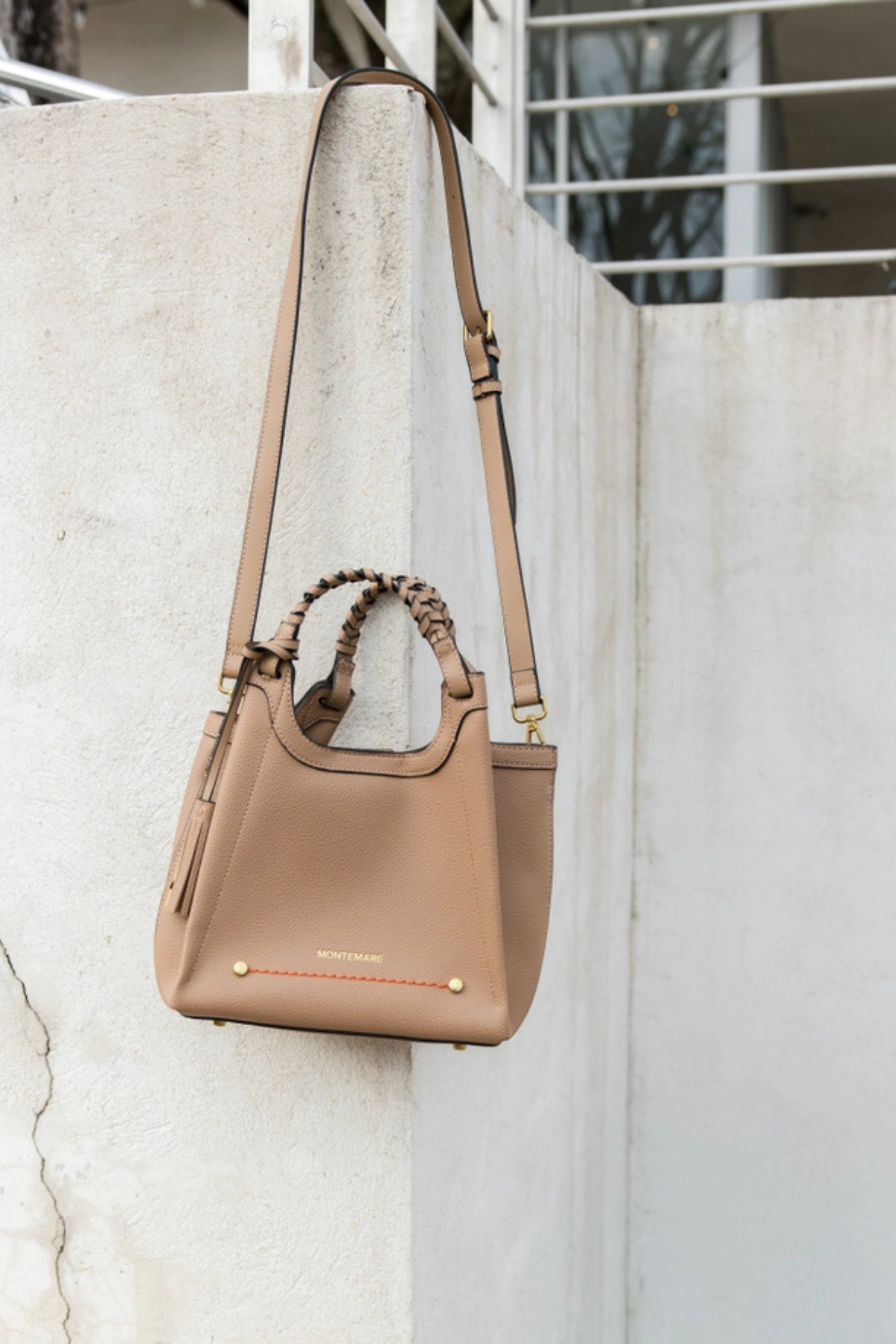 A taupe leather shoulder bag with braided top handles and gold-tone hardware, hanging against a textured concrete wall.