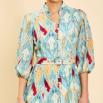 A woman modeling a light blue, yellow, and red ikat-patterned mini dress with puff sleeves and a matching belt with a round buckle, set against a solid beige background.