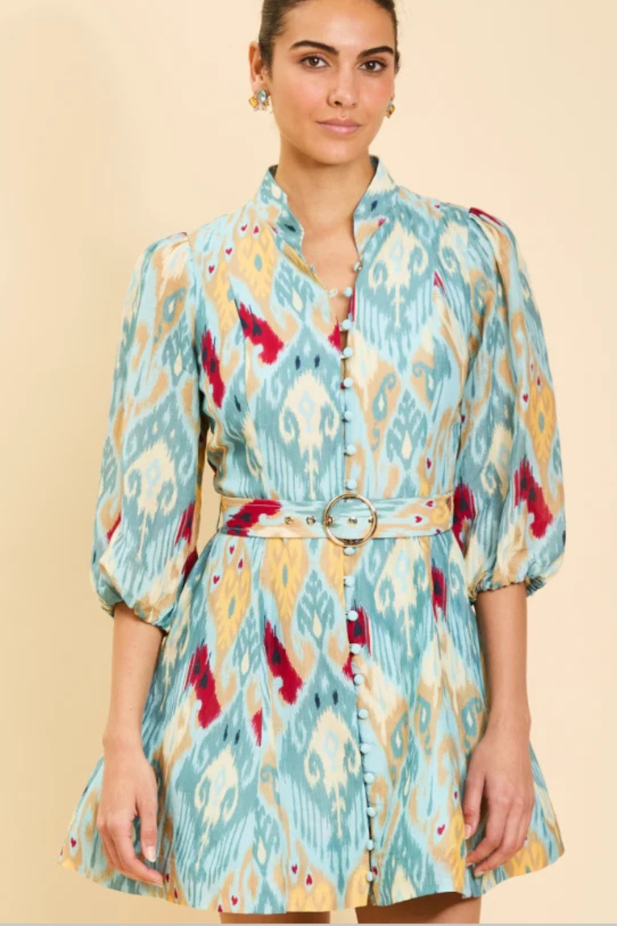 A woman modeling a light blue, yellow, and red ikat-patterned mini dress with puff sleeves and a matching belt with a round buckle, set against a solid beige background.
