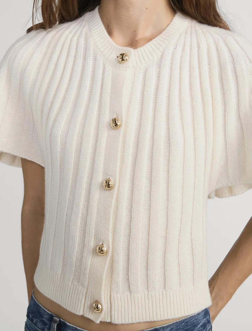 White ribbed knit cardigan with gold buttons worn by a person on a white background