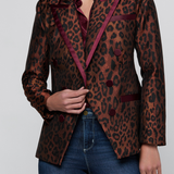 A woman wearing a brown and black leopard-print blazer with deep burgundy satin trim, paired with blue jeans, against a light gray studio background.