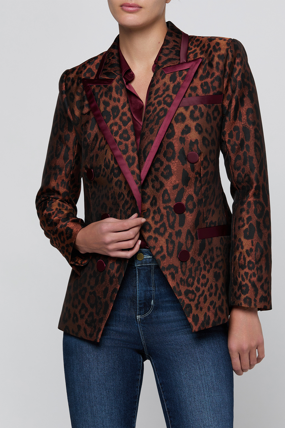 A woman wearing a brown and black leopard-print blazer with deep burgundy satin trim, paired with blue jeans, against a light gray studio background.