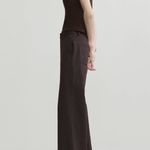 A model shown from the side in a studio setting, wearing a sleeveless dark brown mock-neck top and matching dark brown pinstriped wide-leg trousers with black pointed-toe heels.