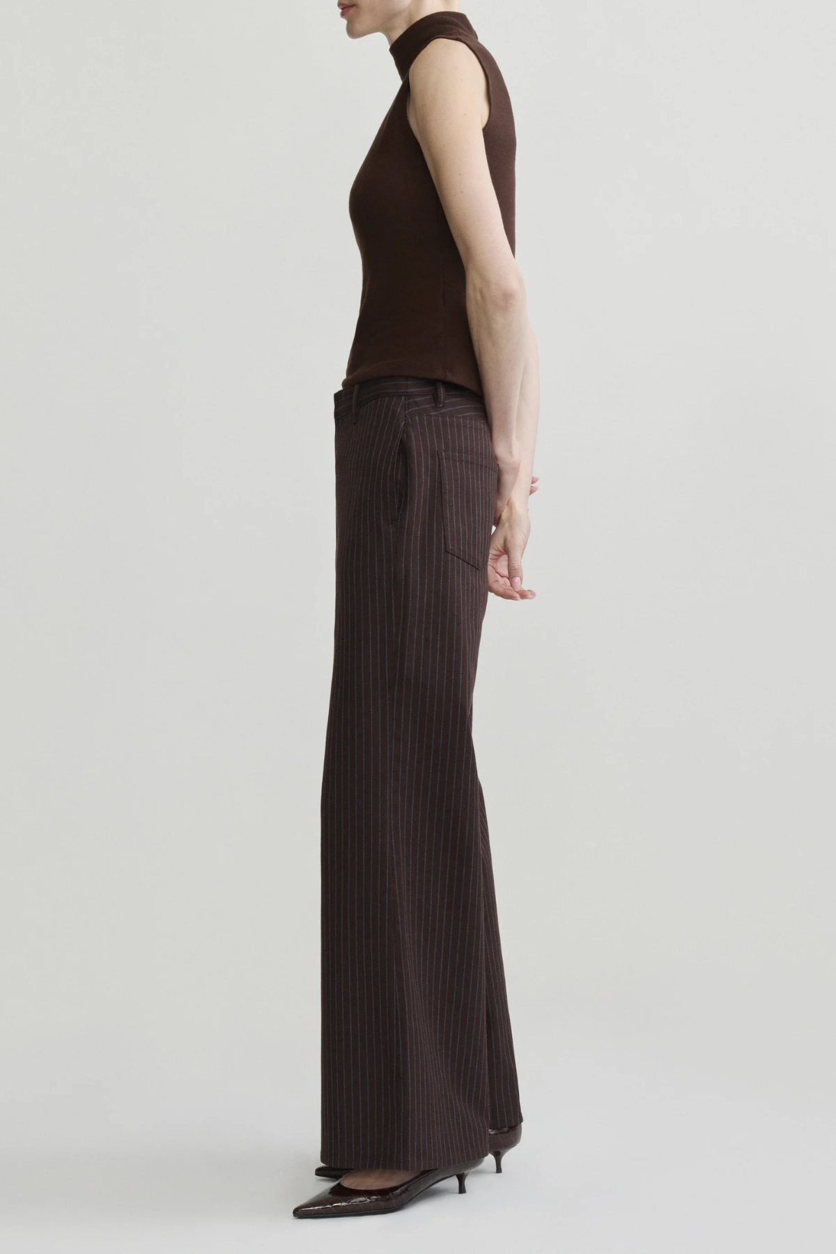 A model shown from the side in a studio setting, wearing a sleeveless dark brown mock-neck top and matching dark brown pinstriped wide-leg trousers with black pointed-toe heels.