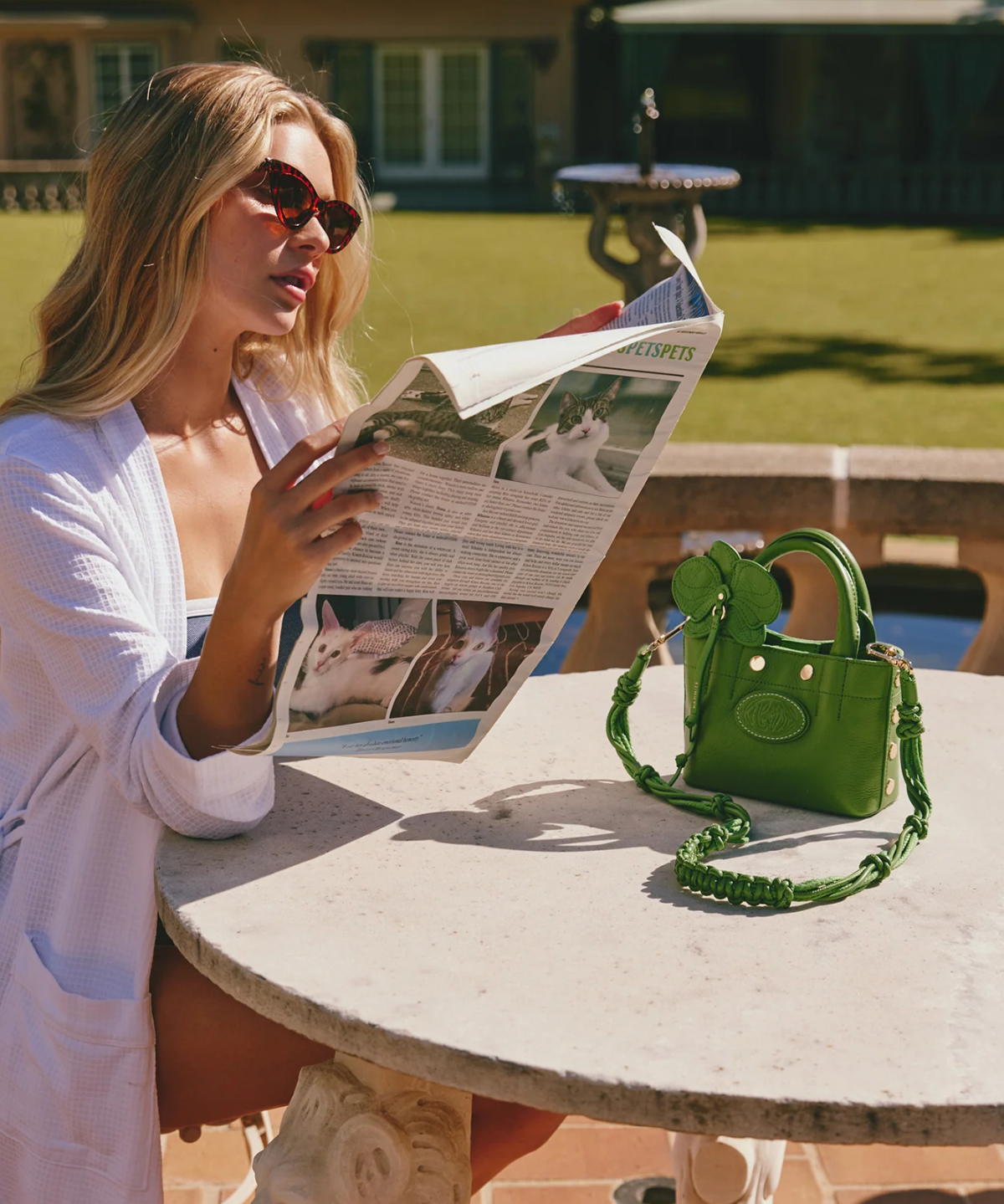 Woman reading a newspaper at an outdoor table with a green handbag beside her.