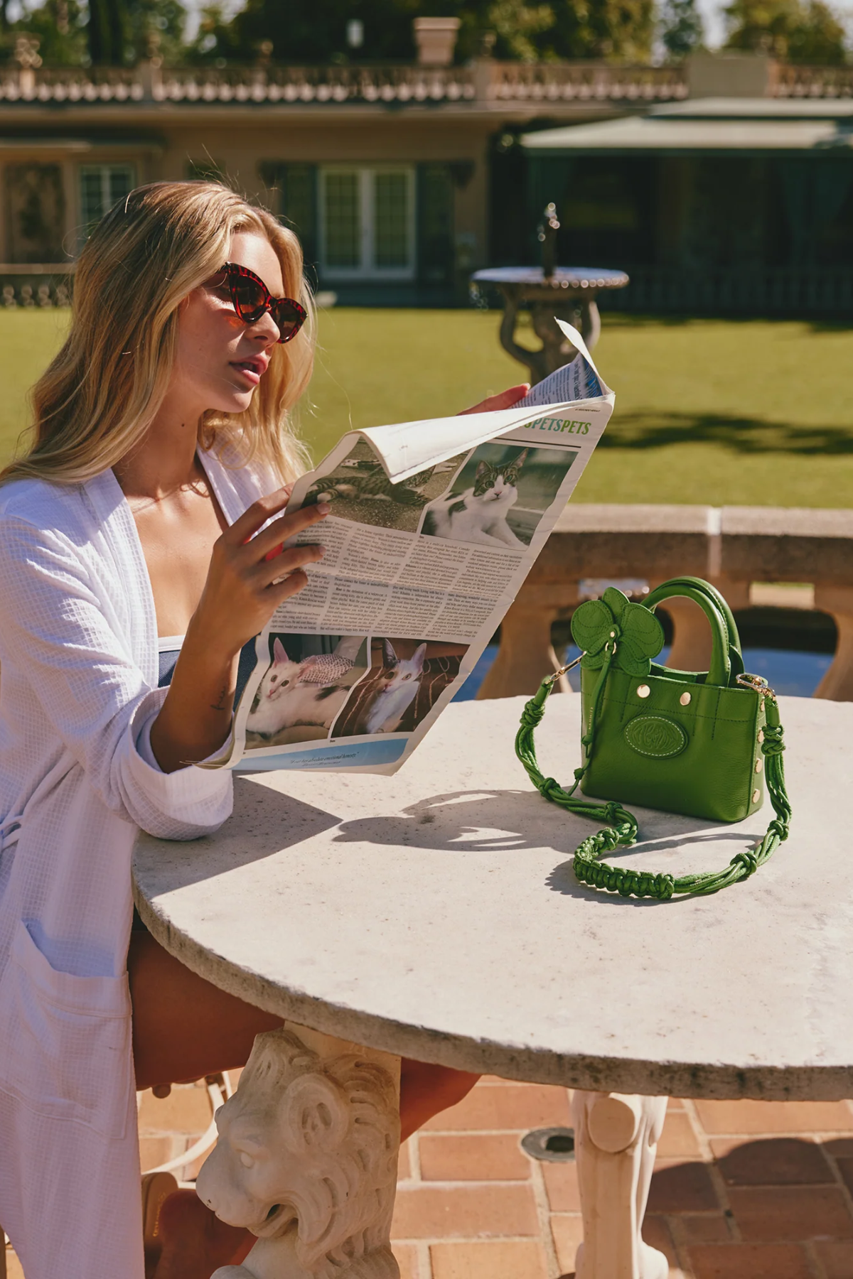 Woman reading a newspaper at an outdoor table with a green handbag beside her.
