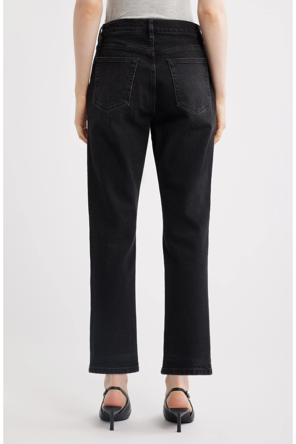 Rear view of a person wearing black cropped jeans with a straight-leg fit, paired with black pointed-toe heels against a light gray studio background.
