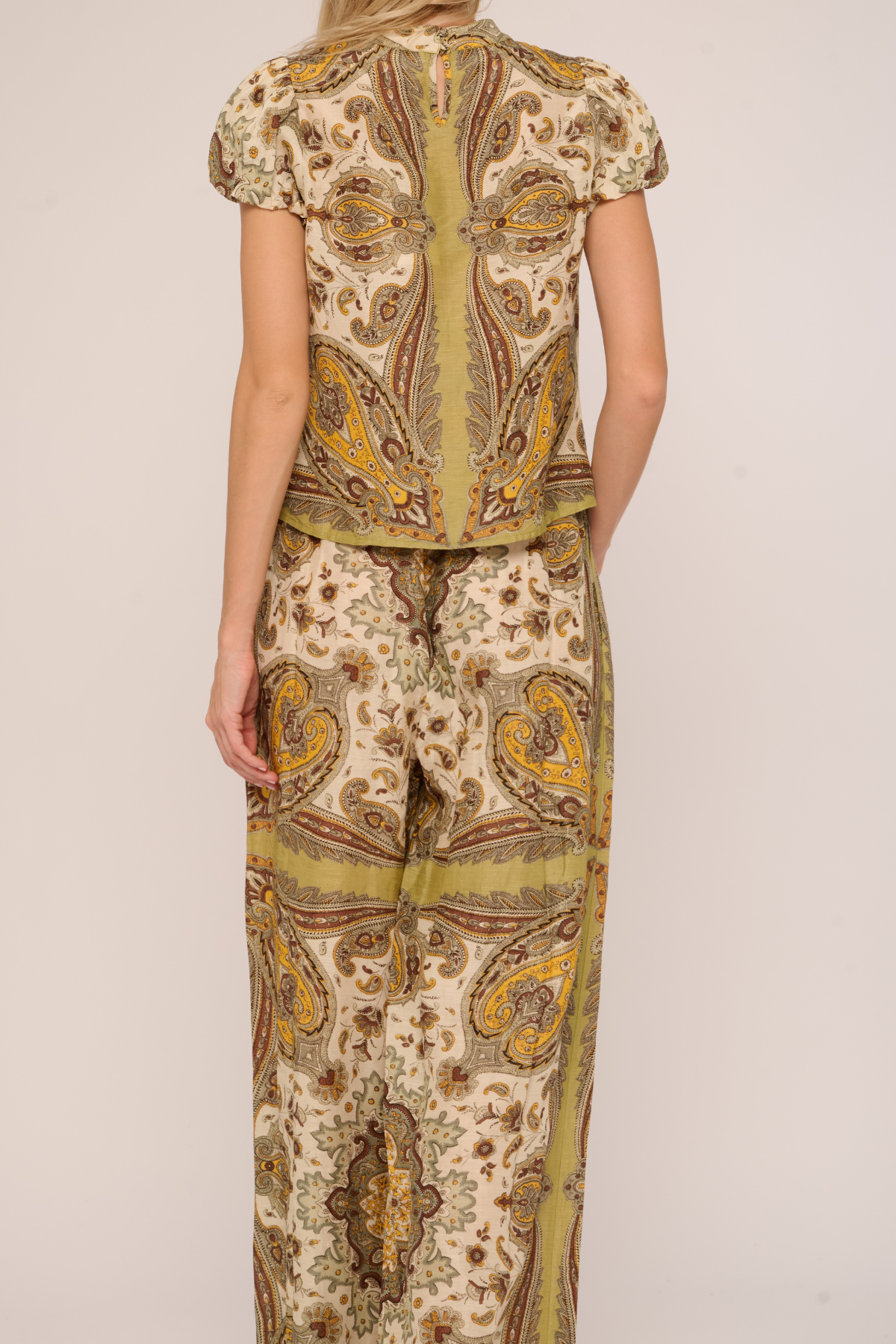 A woman shown from the back wearing a matching two-piece set with a beige, olive green, and brown paisley pattern. The set includes a short-sleeve top and wide-leg pants, photographed against a plain light beige studio background.
