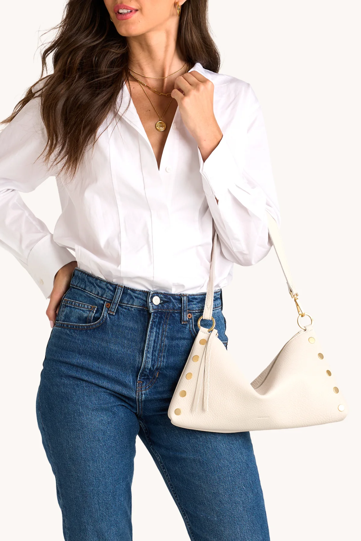 Woman wearing a white shirt and blue jeans holding a cream-colored handbag with gold accents.