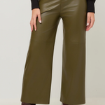 A person wearing olive green wide-leg faux leather pants, styled with a black long-sleeve top and dark brown heeled ankle boots, shown from the waist down against a light gray studio background.
