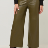 A person wearing olive green wide-leg faux leather pants, styled with a black long-sleeve top and dark brown heeled ankle boots, shown from the waist down against a light gray studio background.
