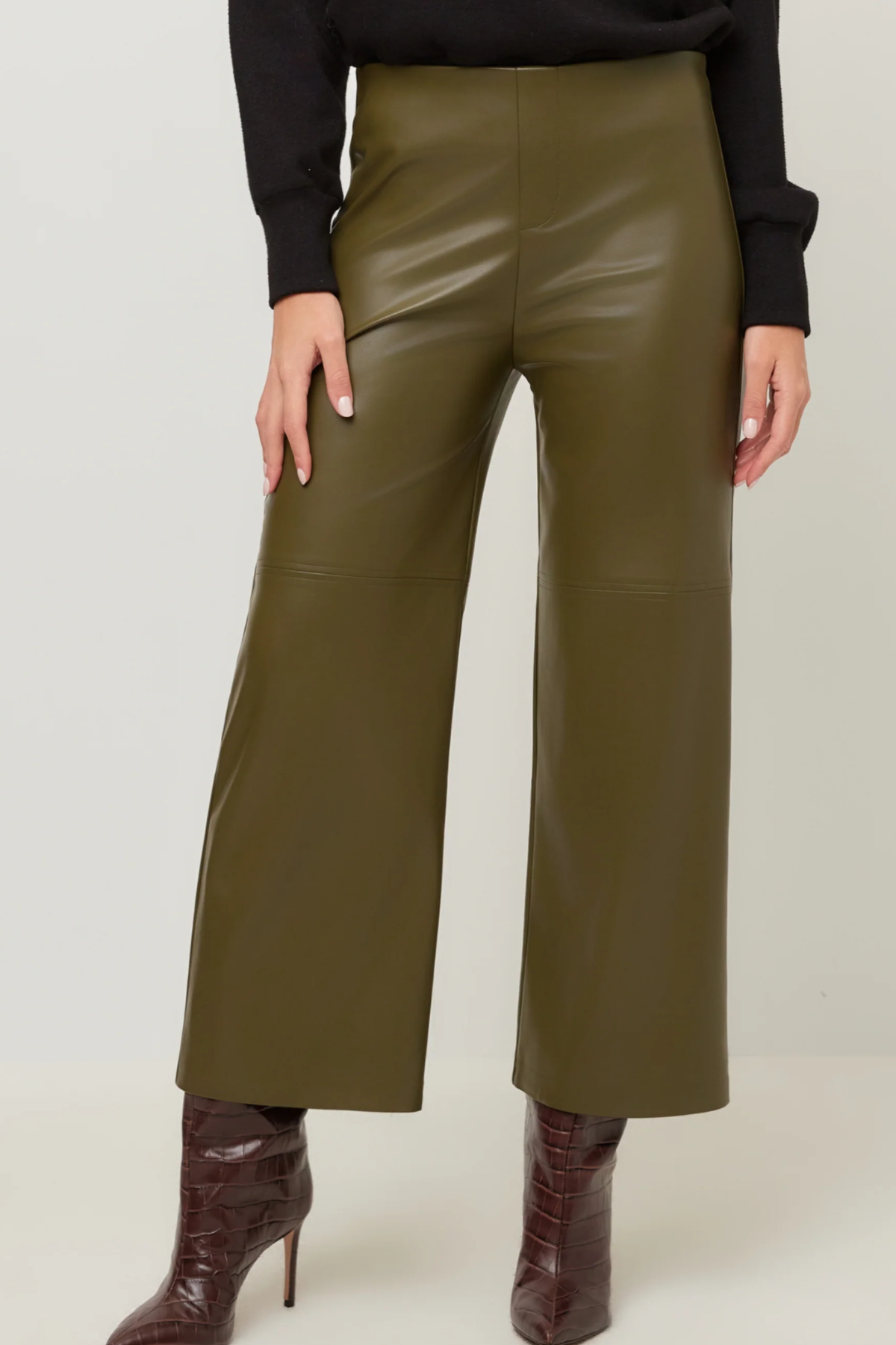 A person wearing olive green wide-leg faux leather pants, styled with a black long-sleeve top and dark brown heeled ankle boots, shown from the waist down against a light gray studio background.