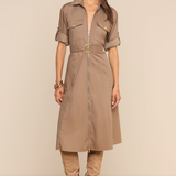 A woman models a taupe-brown short-sleeve shirt dress with a matching belt and gold buckle, styled with matching knee-high boots and sunglasses against a solid beige background.
