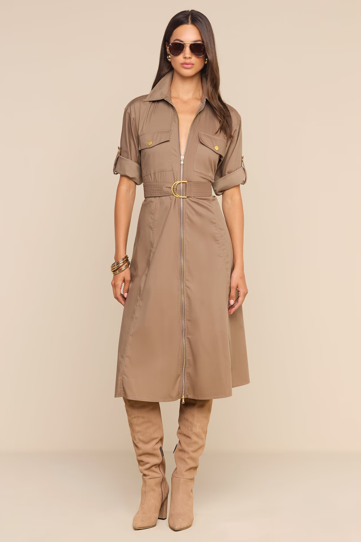A woman models a taupe-brown short-sleeve shirt dress with a matching belt and gold buckle, styled with matching knee-high boots and sunglasses against a solid beige background.