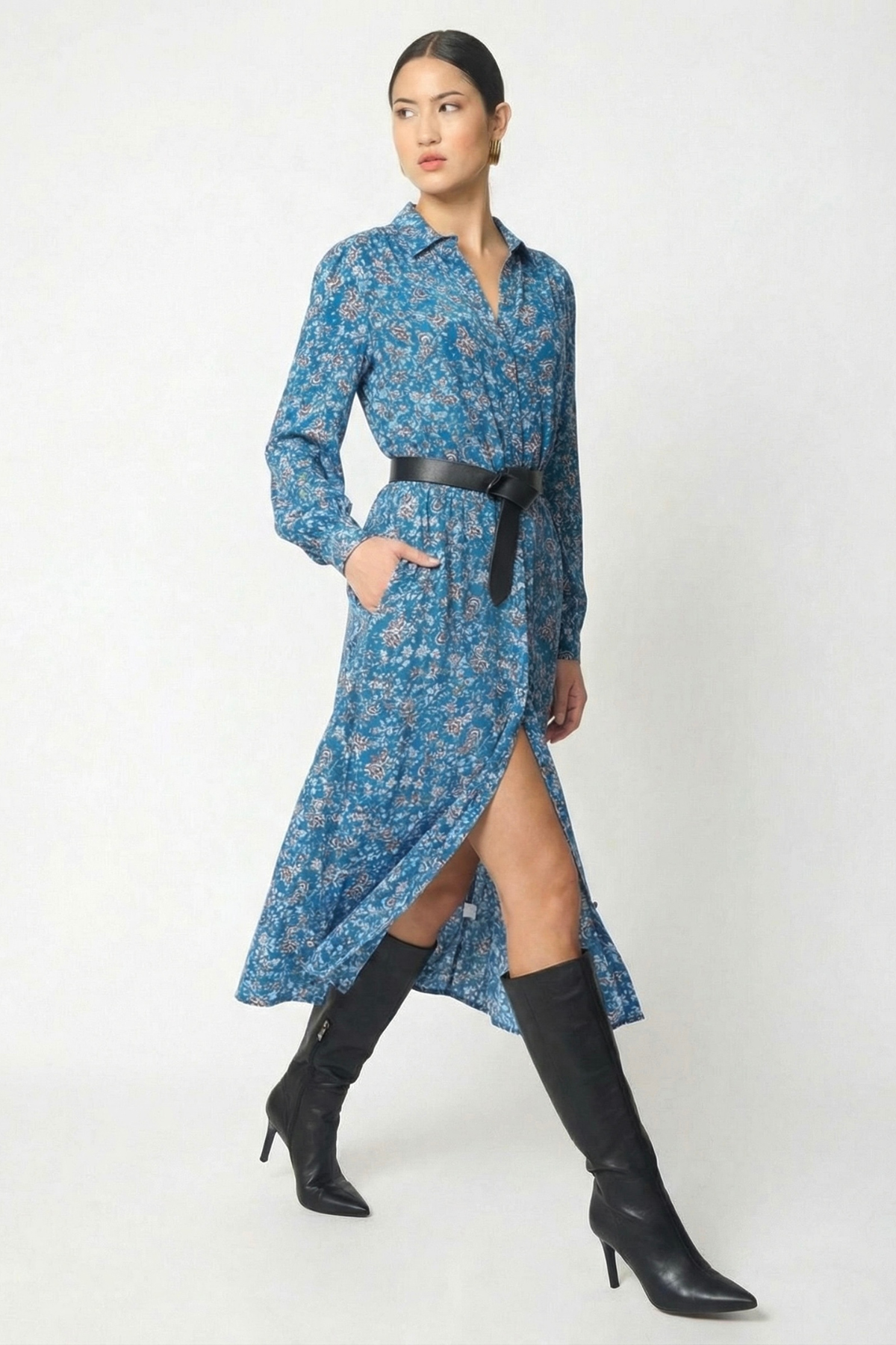 A woman models a blue floral long-sleeve midi dress with a black belt and black knee-high heeled boots against a plain light gray studio background.