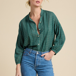 A woman modeling a green plaid button-up shirt with blue jeans against a solid beige background.
