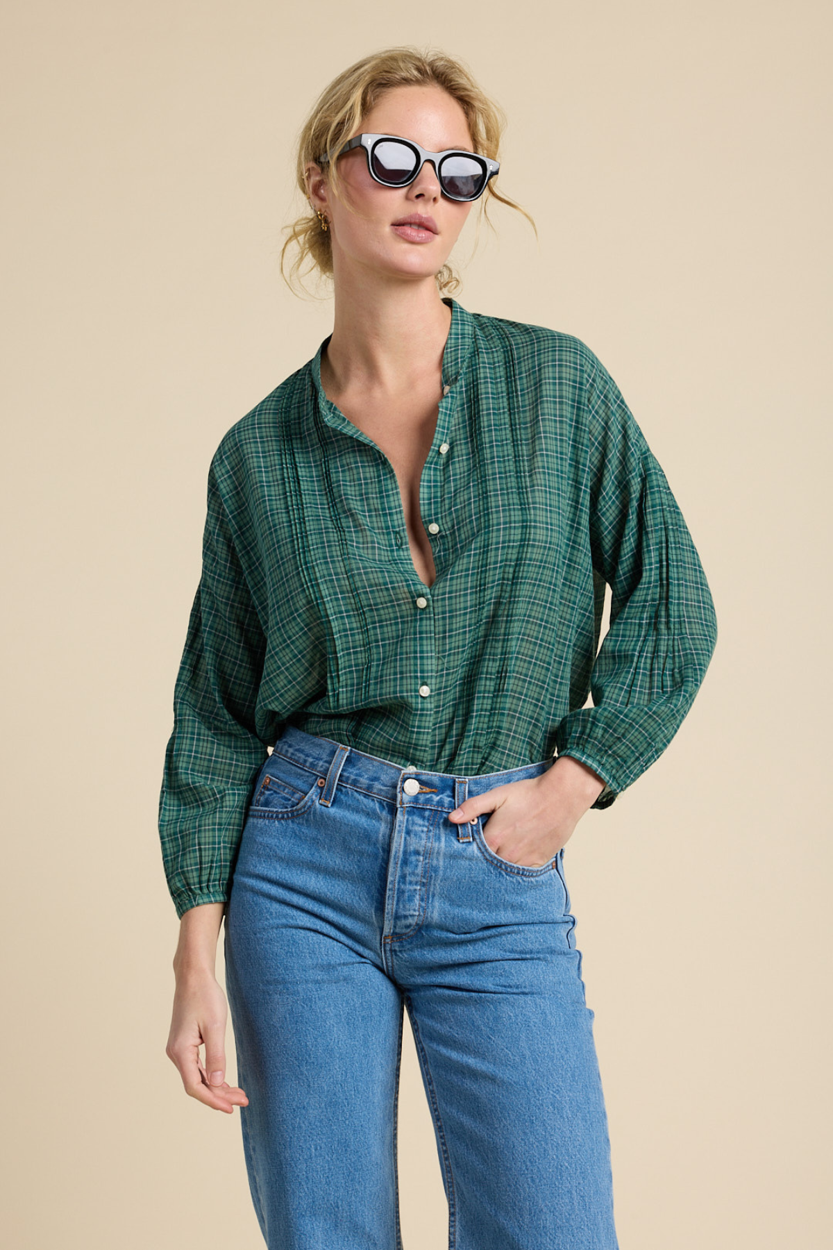 A woman modeling a green plaid button-up shirt with blue jeans against a solid beige background.