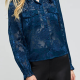 A woman wearing a dark blue sheer floral lace long-sleeve blouse with button details and black pants, photographed against a light gray studio background.