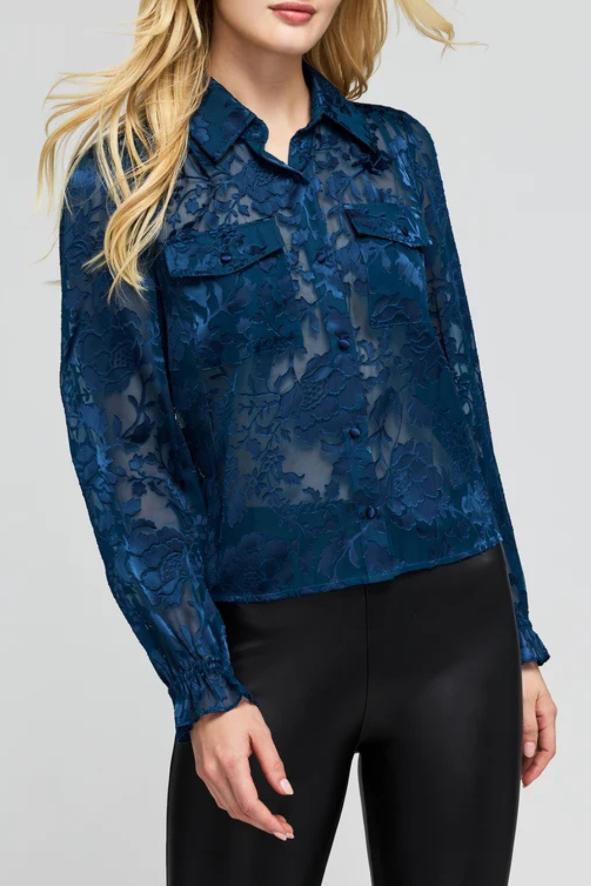 A woman wearing a dark blue sheer floral lace long-sleeve blouse with button details and black pants, photographed against a light gray studio background.