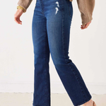 A person wearing dark blue cropped flare jeans with whiskering and distressed details on the thigh, styled with a beige sweater and brown and black pointed-toe slingback flats.