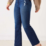 A person wearing dark blue cropped flare jeans with whiskering and distressed details on the thigh, styled with a beige sweater and brown and black pointed-toe slingback flats.