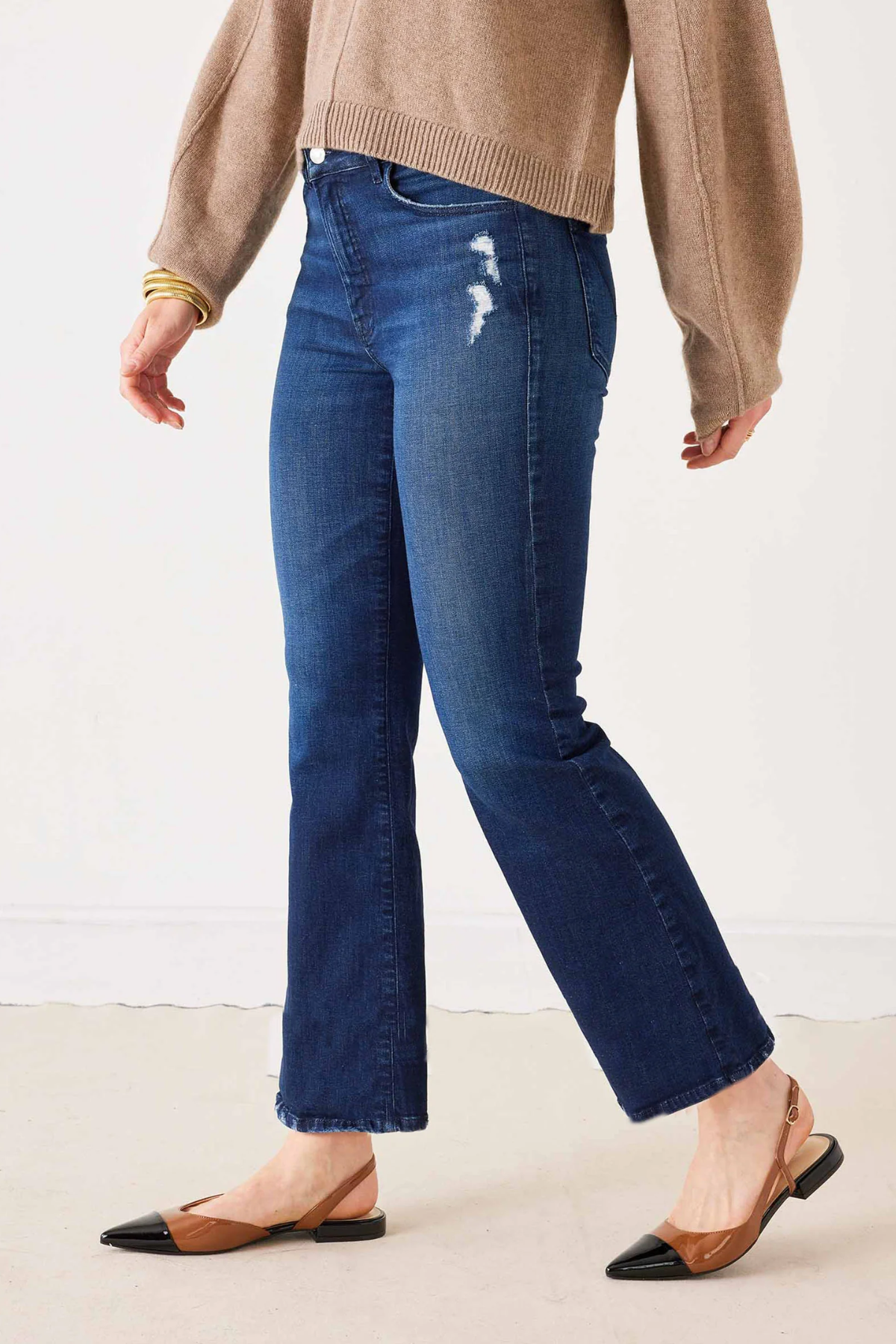 A person wearing dark blue cropped flare jeans with whiskering and distressed details on the thigh, styled with a beige sweater and brown and black pointed-toe slingback flats.
